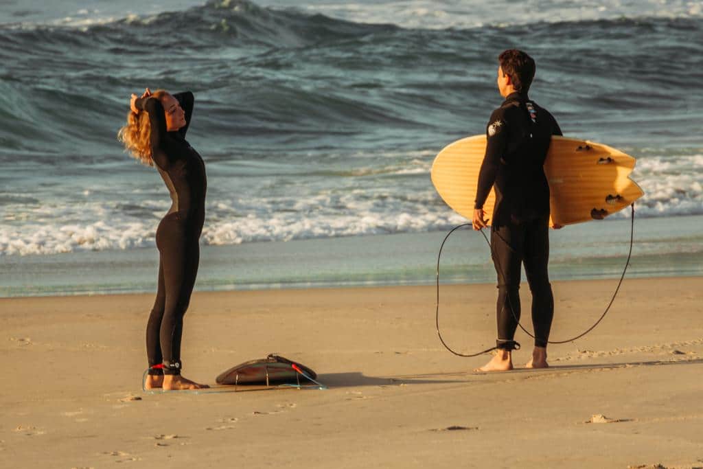 casal surfista, com roupas de surf e prancha, se preparando para entrar na água.